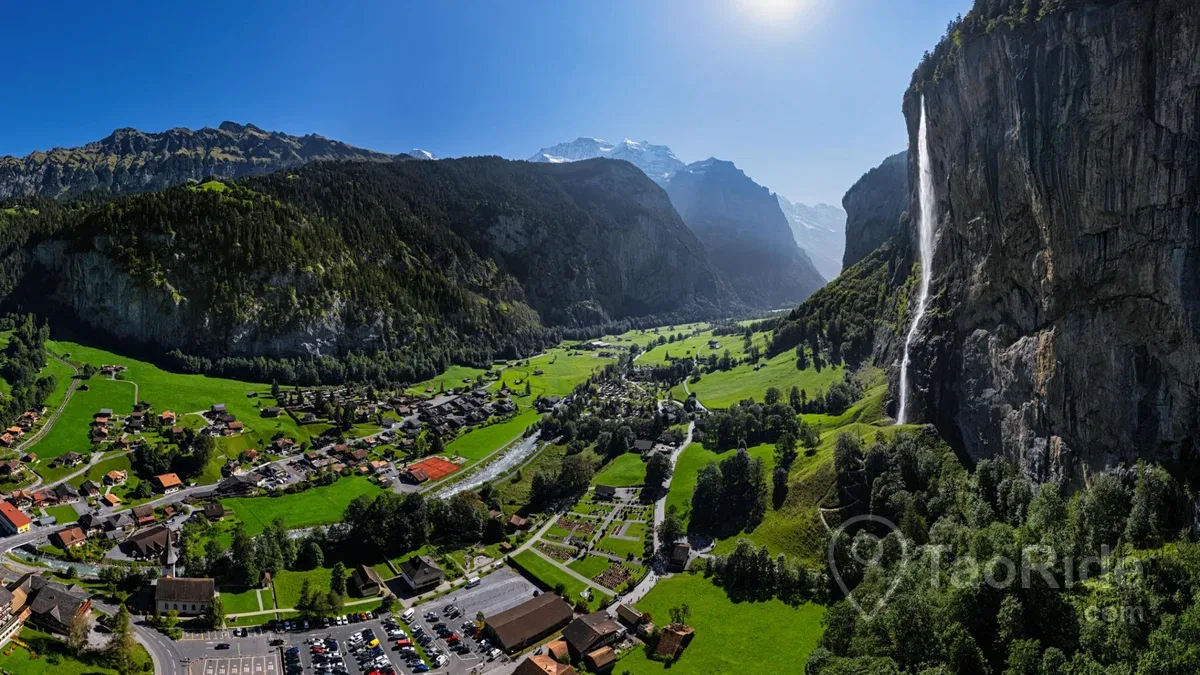 Panoramic view of Lauterbrunnen Valley in Switzerland with Staubbach Falls, traditional Swiss chalets, green meadows, and towering Alpine cliffs.