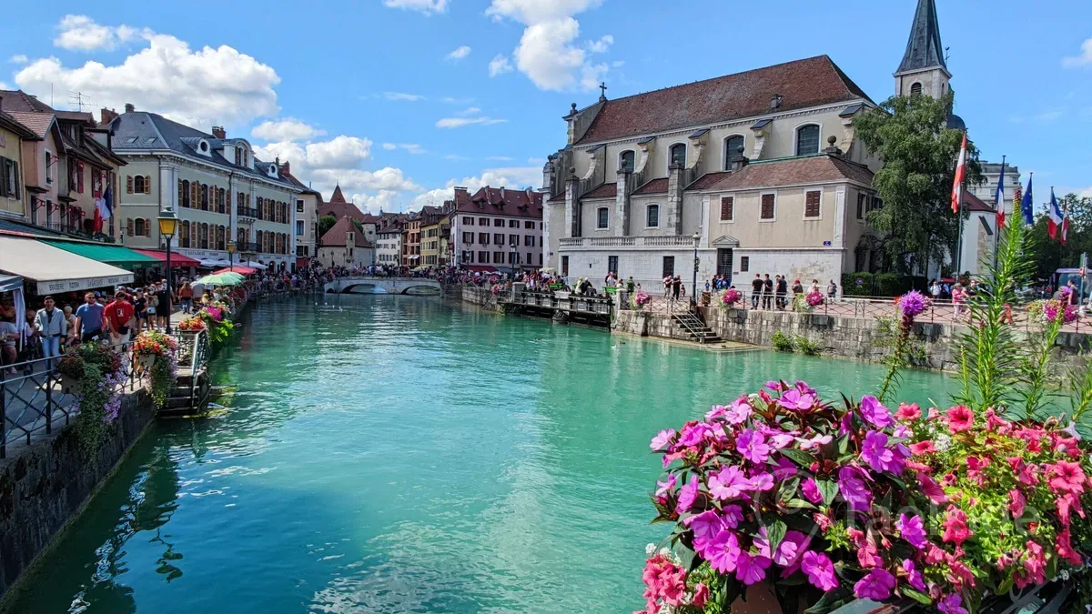 Beautiful Annecy, France, featuring the historic Old Town, colorful canals, flower-lined bridges, Lake Annecy, and the surrounding French Alps on a sunny summer day.
